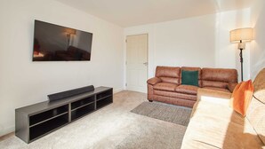 A living room with a flat-screen TV mounted on the wall, a brown leather sofa, a small black entertainment unit, and a floor lamp. - Host Stay Aronora Guest House (Blaydon on Tyne)