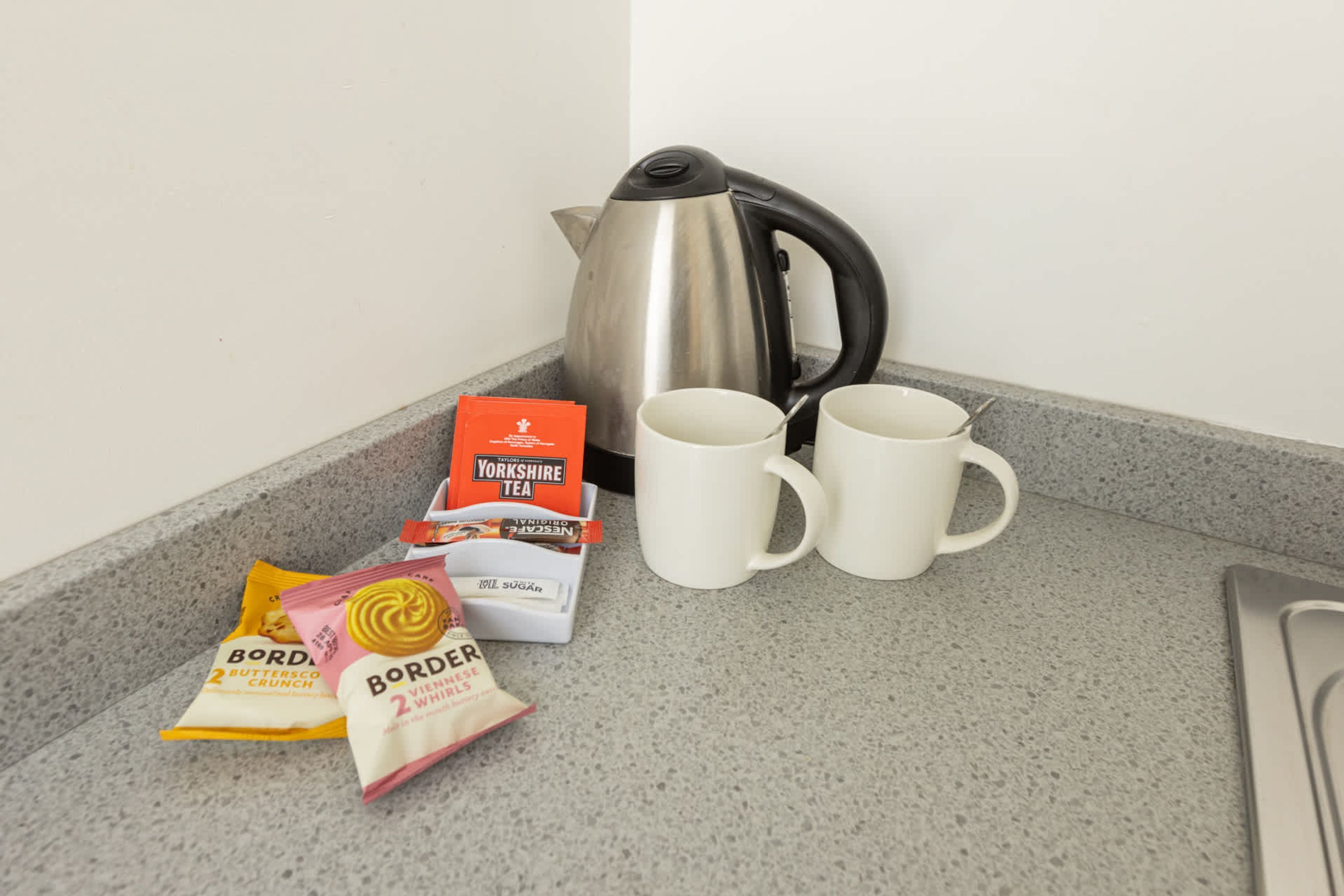 A kitchen counter with a kettle, two white mugs, and packets of tea and biscuits.