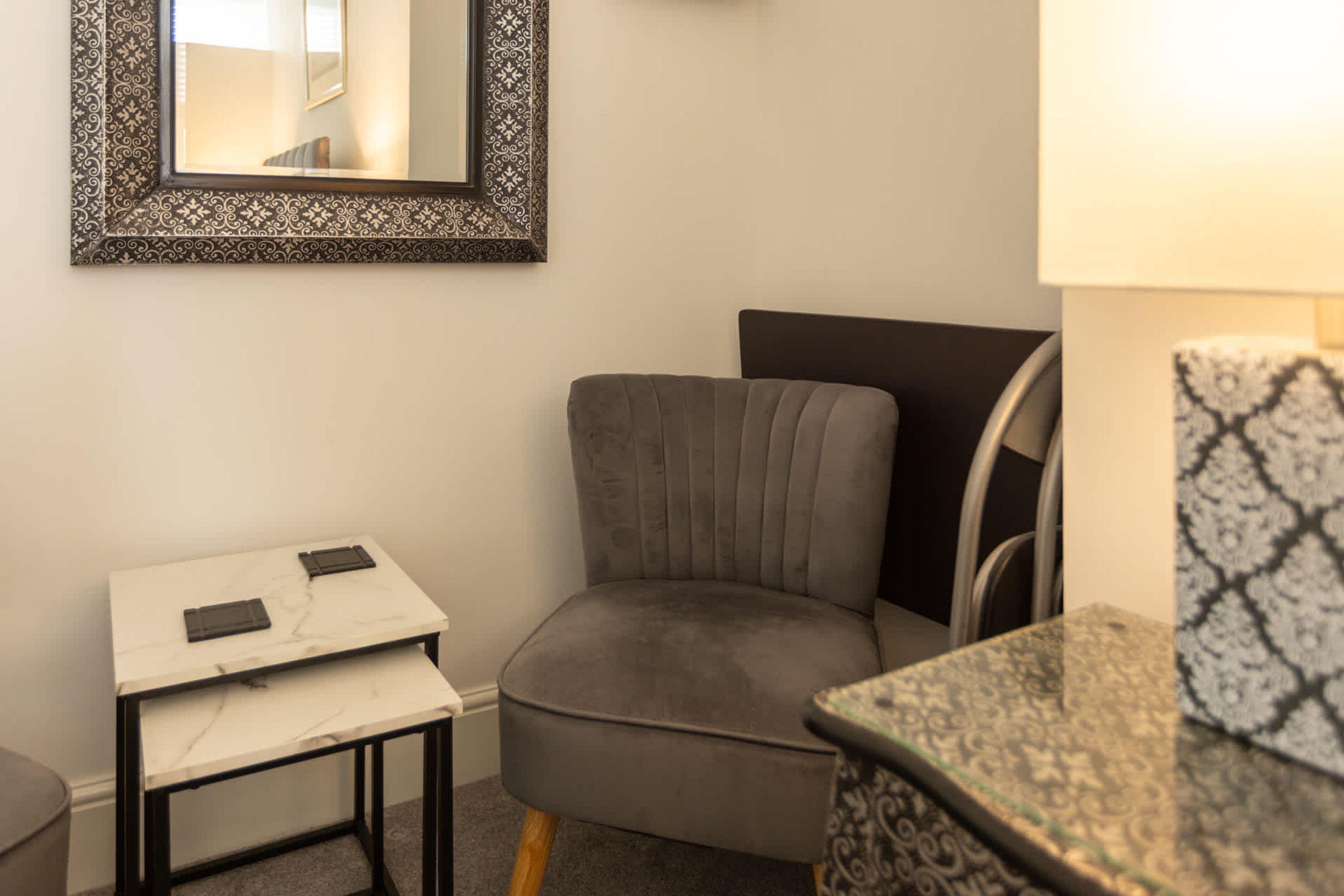 A room with a grey upholstered chair, a small white table with a marble top, and a patterned side table.