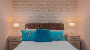 A bedroom with a wooden headboard, two bedside tables with lamps, a bed with floral bedding, and a fluffy blue pillow. - Host Stay Pied-a-terre at Manor View (Bury)