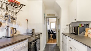 A compact kitchen with a washing machine, a kettle, and a toaster. A 'WELCOME' sign is visible in the hallway. - Host Stay Fishermans Cottage (Cromer)