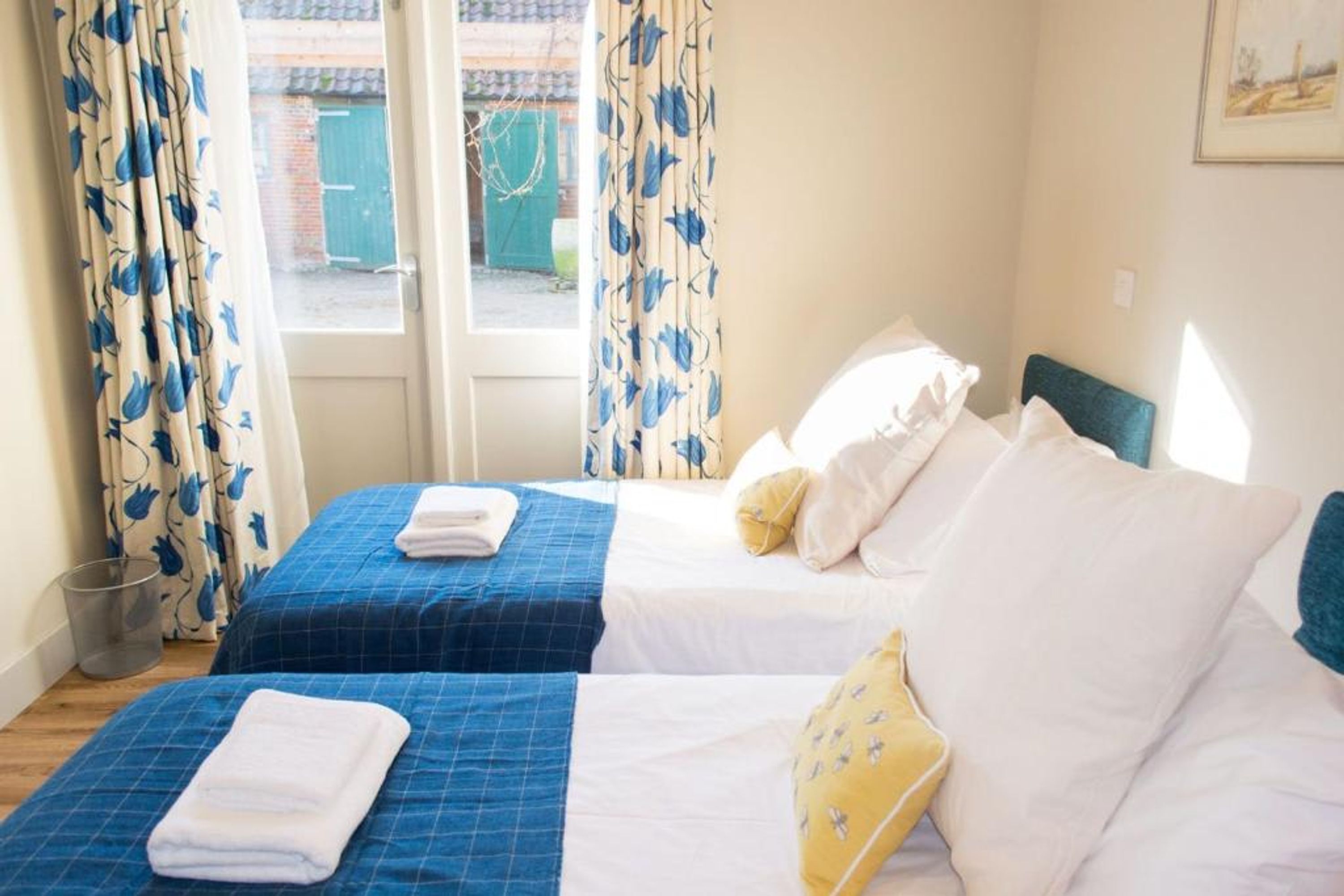 A hotel room with two beds, each with a blue bedspread and white linens. There are curtains with a blue floral pattern, and a window with a view of a building outside.