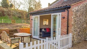 A cozy outdoor seating area with a table and chairs, a white picket fence, and a brick building with a glass door leading to a room with a sofa and a window. - Host Stay Boathouse Barn (Norwich)