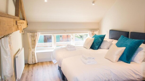 A bedroom with two beds, a window with curtains, wooden beams on the ceiling, and a radiator. - Host Stay Boathouse Barn (Norwich)