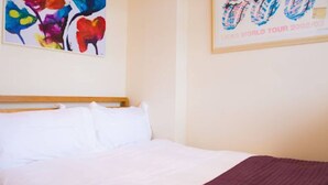 Ett hotellrum med en säng, vita sängkläder, en färgstark, abstrakt målning och en inramad affisch på väggen.