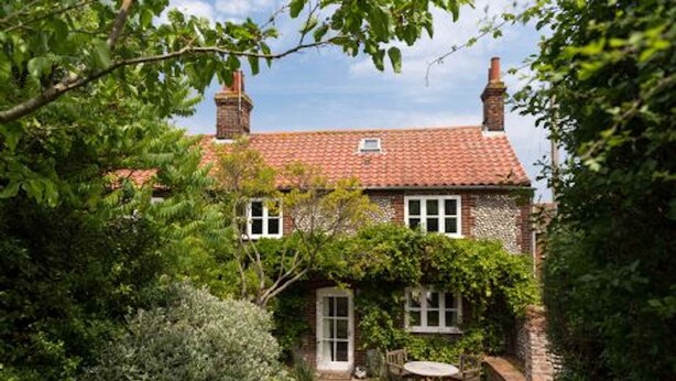 A traditional brick house with a red-tiled roof, surrounded by lush greenery and a small garden with a table and chairs.