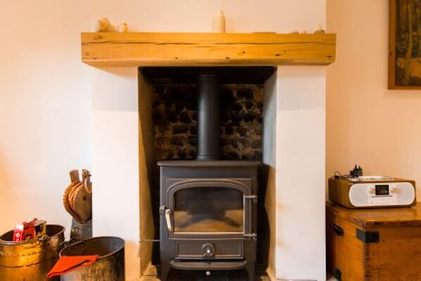 A wood-burning stove with a chimney, a wooden shelf above it, and a small heater on a cabinet to the right. - Host Stay Marlpit Cottage (Holt)