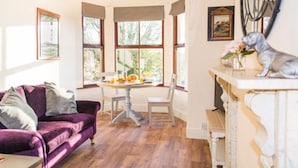 A cozy dining area with a purple sofa, a round table with orange fruit, and a white fireplace with a dog figurine.