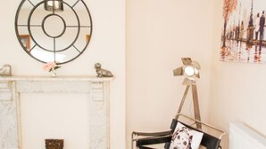 A room with a fireplace, a round mirror, a tripod lamp, and a chair with a cushion.