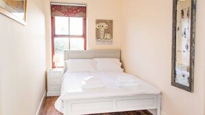 A bedroom with a white bed, a nightstand, a window with red curtains, and a mirror on the wall.