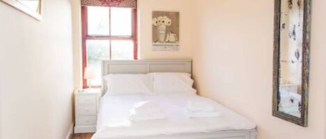 A bedroom with a white bed, a nightstand, a window with red curtains, and a mirror on the wall.