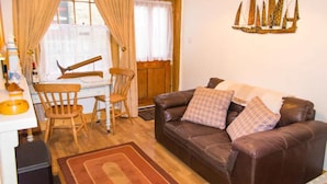 Un salon avec un canapé en cuir marron, deux coussins à carreaux, une table à manger en bois avec des chaises, un meuble en bois et une fenêtre avec des rideaux.