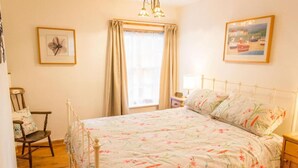 Une chambre à coucher avec une couvre-lit à motifs floraux, une chaise en bois, une table de chevet avec une lampe et une fenêtre avec des rideaux.