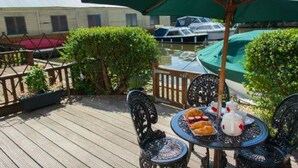 A wooden deck with a table set for a meal, surrounded by greenery and overlooking a body of water with boats. - Host Stay Quayside Cottage (Norwich)