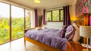 A bedroom with a large window, a bed with a purple blanket, and a view of greenery outside. - Host Stay Four Seasons (Norwich)