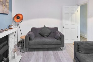 A living room with a grey sofa, a floor lamp, a white fireplace, and a white door. - Host Stay Thurstane Street (Bolton)