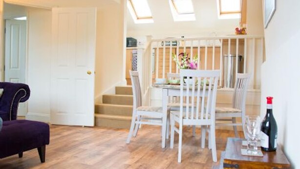 Una habitación con piso de madera, un sofá púrpura, una mesa de comedor blanca con sillas y una escalera que conduce a un nivel superior.