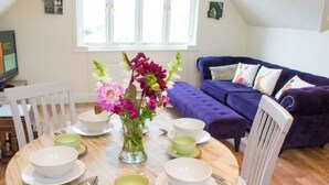 Un comedor con una mesa redonda de madera dispuesta para cuatro personas, rodeada de sillas blancas. Un jarrón con flores se encuentra sobre la mesa.