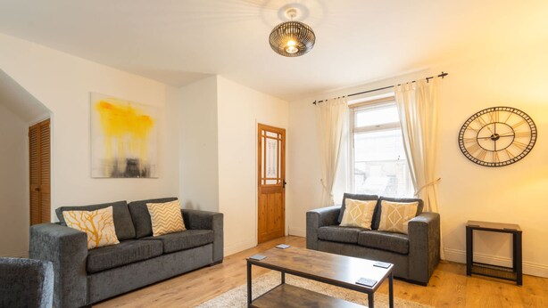 A modern living room with two grey sofas, a wooden coffee table, and a large wall clock. - Host Stay Wind Rose Cottage (Morpeth)