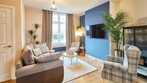 Un salón con sofá, sillón y una mesa de centro. Hay un televisor montado en la pared, una planta en maceta y una ventana con cortinas.