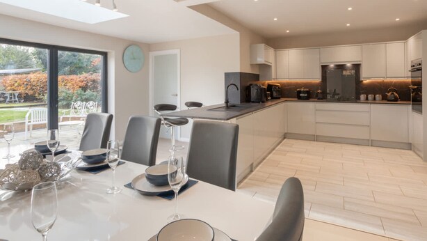 A modern kitchen with a central island, a dining area with a table set for four, and a large window with a view of a garden. - Host Stay High Gables (Northampton)