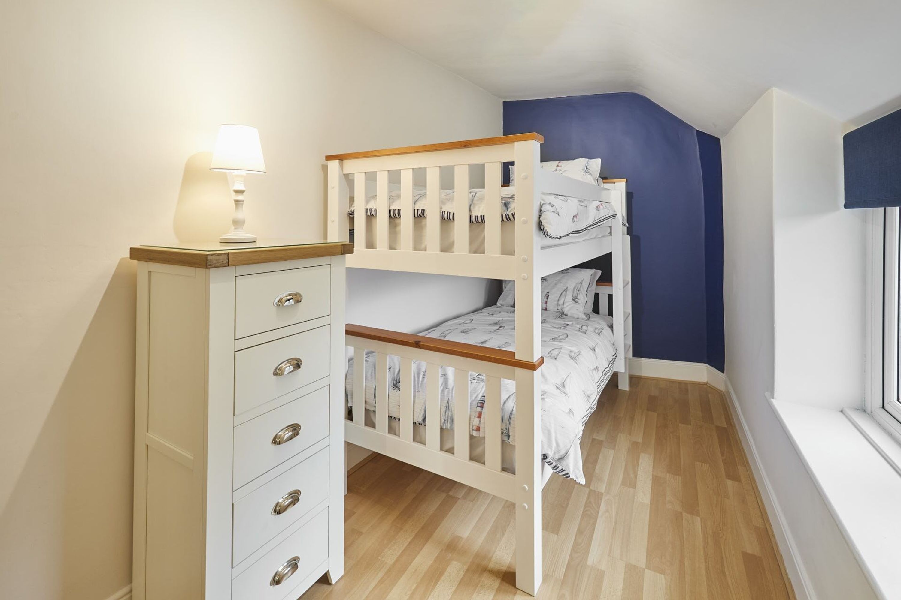 A bunk bed with a wooden frame and white bedding, a white dresser with drawers, and a lamp on top.