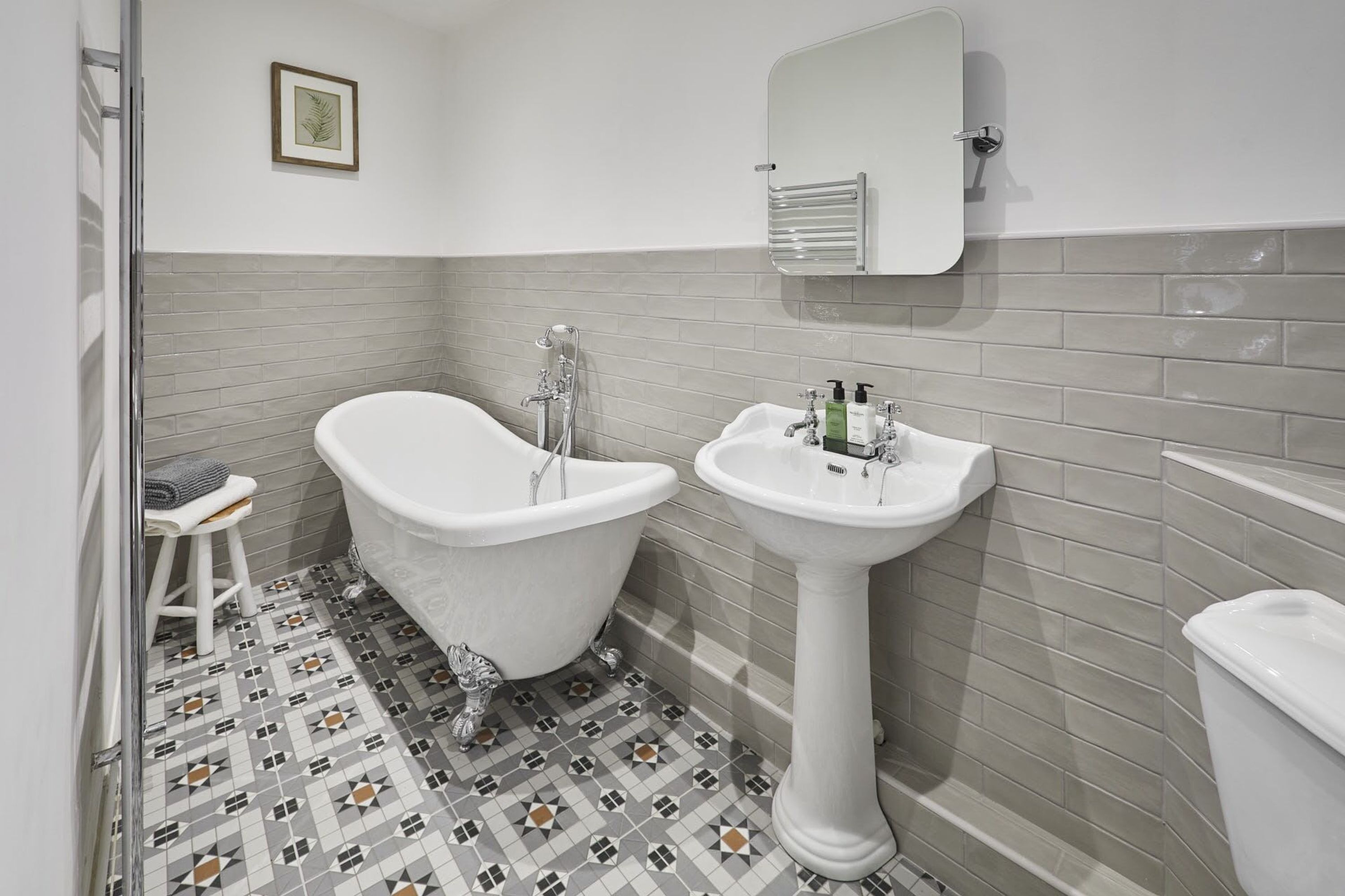 A bathroom with a bathtub, a pedestal sink, and a toilet. There is a small stool and a framed picture on the wall.