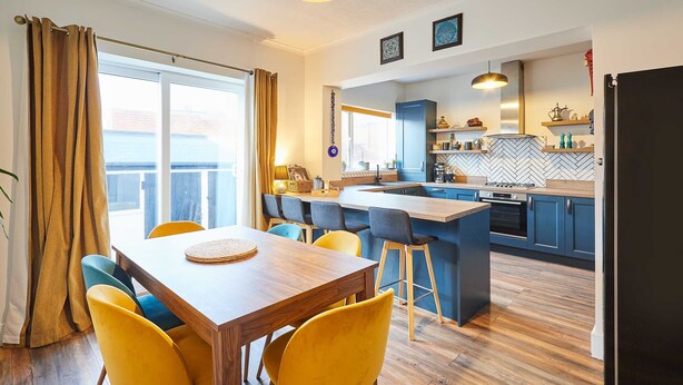 A modern kitchen with a central island, wooden dining table with yellow chairs, and a view of the outside through large windows.