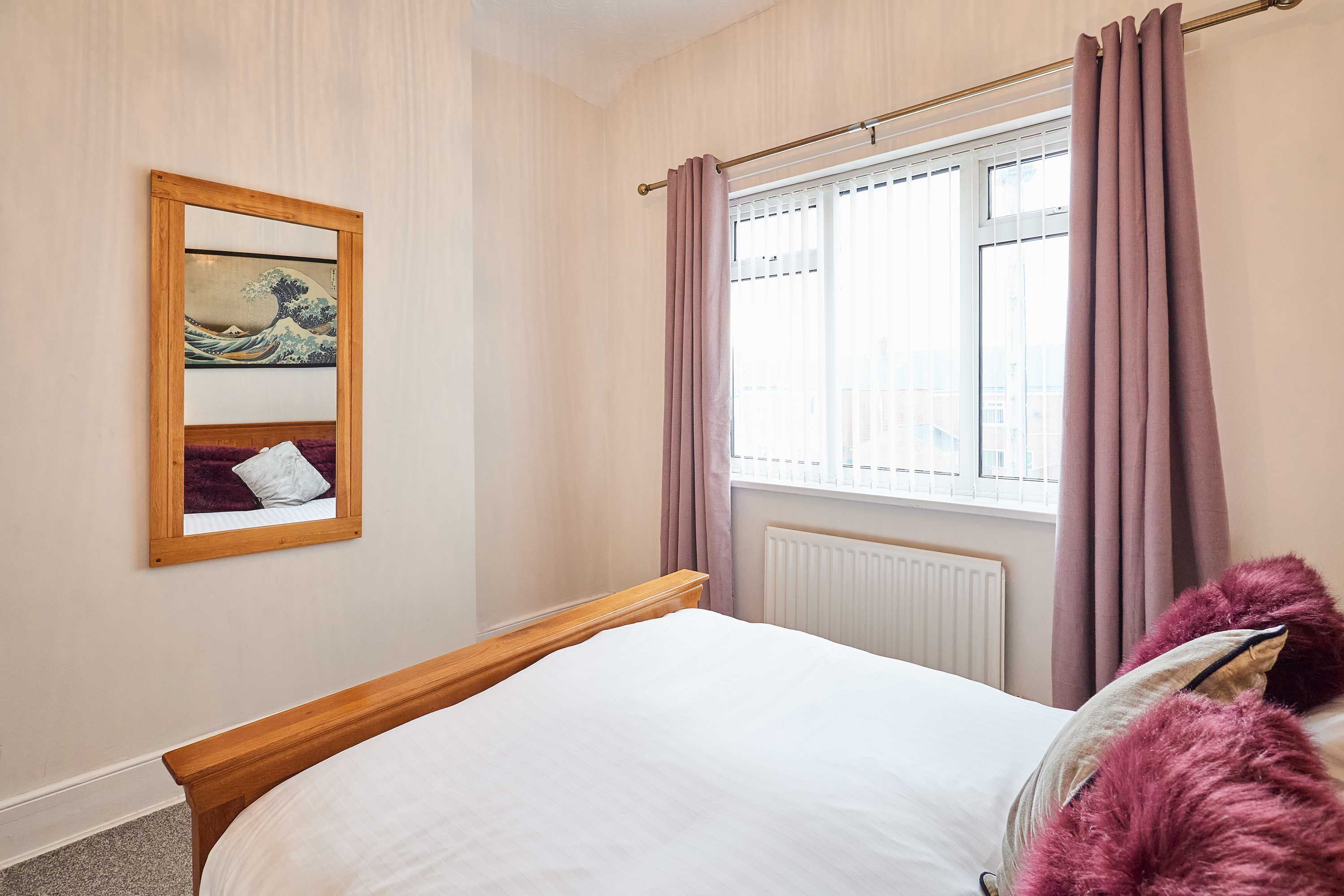 A bedroom with a wooden bed, a window with curtains, a radiator, and a framed artwork on the wall.