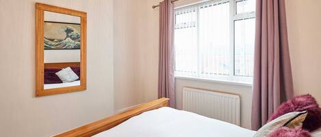 A bedroom with a wooden bed, a window with curtains, a radiator, and a framed artwork on the wall.