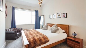 A bedroom with a large bed, a wooden nightstand, a leather armchair, a mirror, and a chandelier.