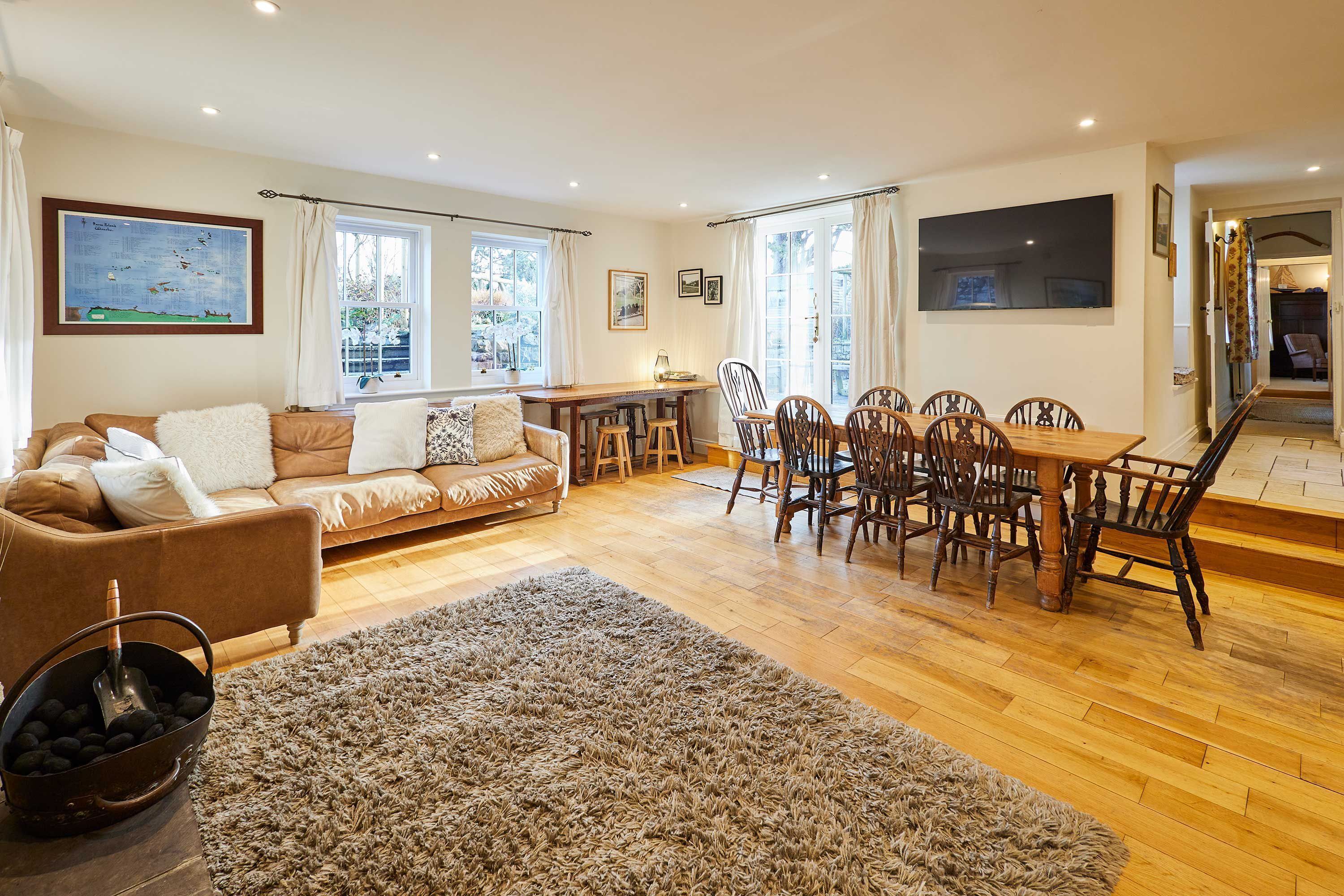A spacious living area with a wooden floor, a brown sofa, a dining table with chairs, and a large window with curtains.