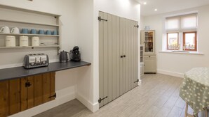 A kitchen with a toaster, coffee maker, and shelves with kitchenware. There is a door with a handle and a window with blinds. - Host Stay Spittal Farm (Wigton)