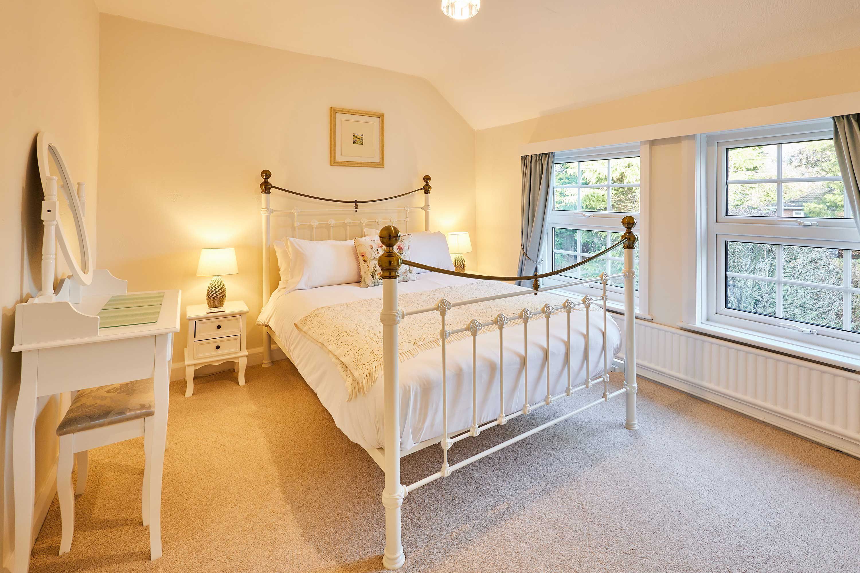 A neatly arranged bedroom with a four-poster bed, a bedside table with a lamp, a small desk with a mirror, and a window with curtains.