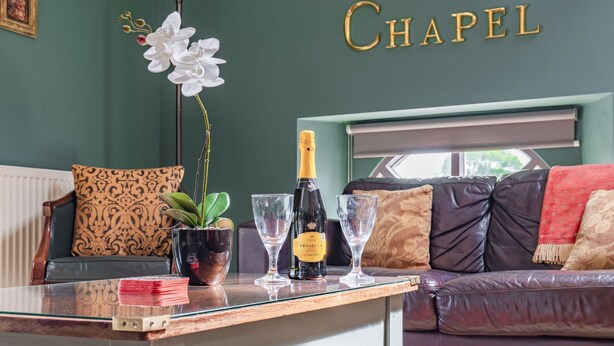 A living room with a glass table, a bottle of champagne, and two glasses. There is a sofa with patterned cushions and a potted plant. - Host Stay Nine Ladies (Matlock)