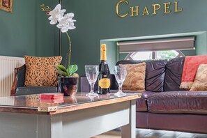 A living room with a glass table, a bottle of champagne, and two glasses. There is a sofa with patterned cushions and a potted plant. - Host Stay Nine Ladies (Matlock)