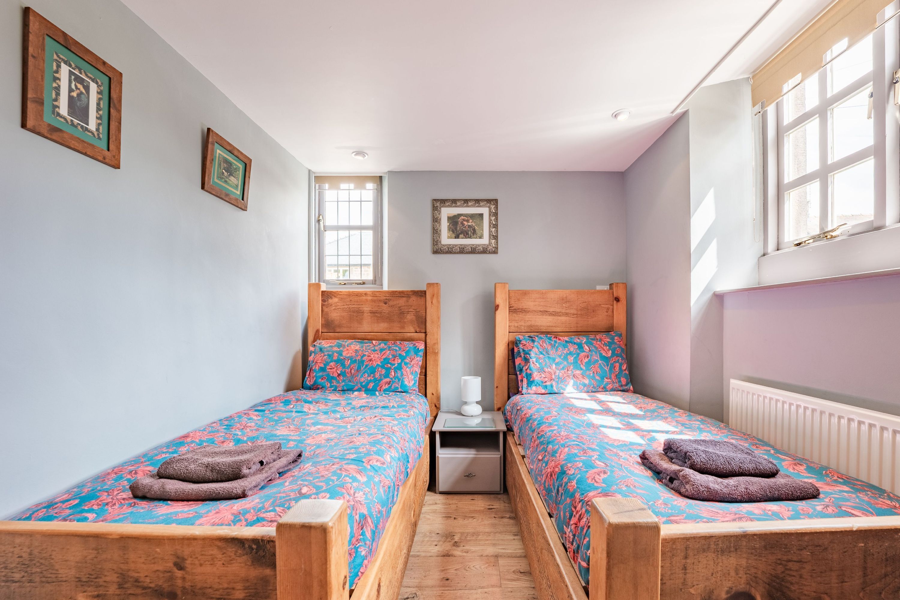 Ein Doppelzimmer mit zwei hölzernen Betten, floralem Bettzeug, einem Nachttisch und einem Fenster mit Jalousien.