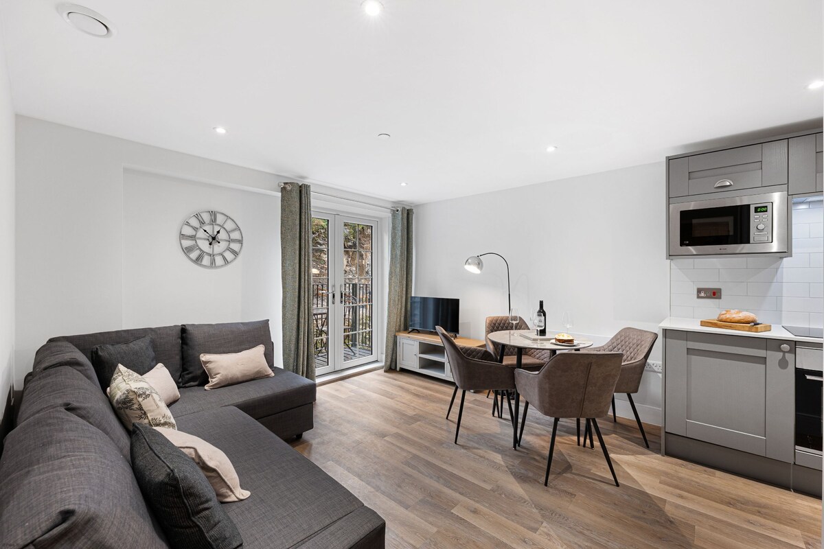 A modern living room with a grey sofa, a dining area with a round table and chairs, and a kitchen with a microwave and cabinets.
