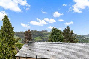 A view of a house with a stone chimney and a tiled roof, overlooking a green landscape and a castle on a hill. - Host Stay 44 Wellington Street (Matlock)