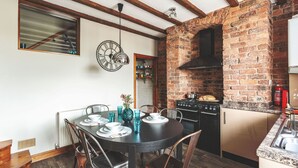 A dining area with a round table set for four, surrounded by wooden chairs. A brick wall oven is visible in the background, and a window with blinds is on the left. - Host Stay 44 Wellington Street (Matlock)