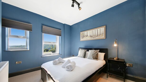 Un dormitorio con una cama grande, dos ventanas, un cuadro, una mesita de noche con lámpara y vistas al exterior.