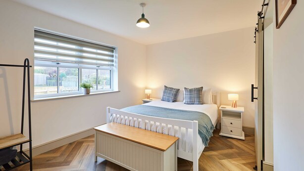 Une chambre à coucher comprenant un lit, des tables de chevet, un banc, un radiateur, une fenêtre avec des stores et un luminaire suspendu.