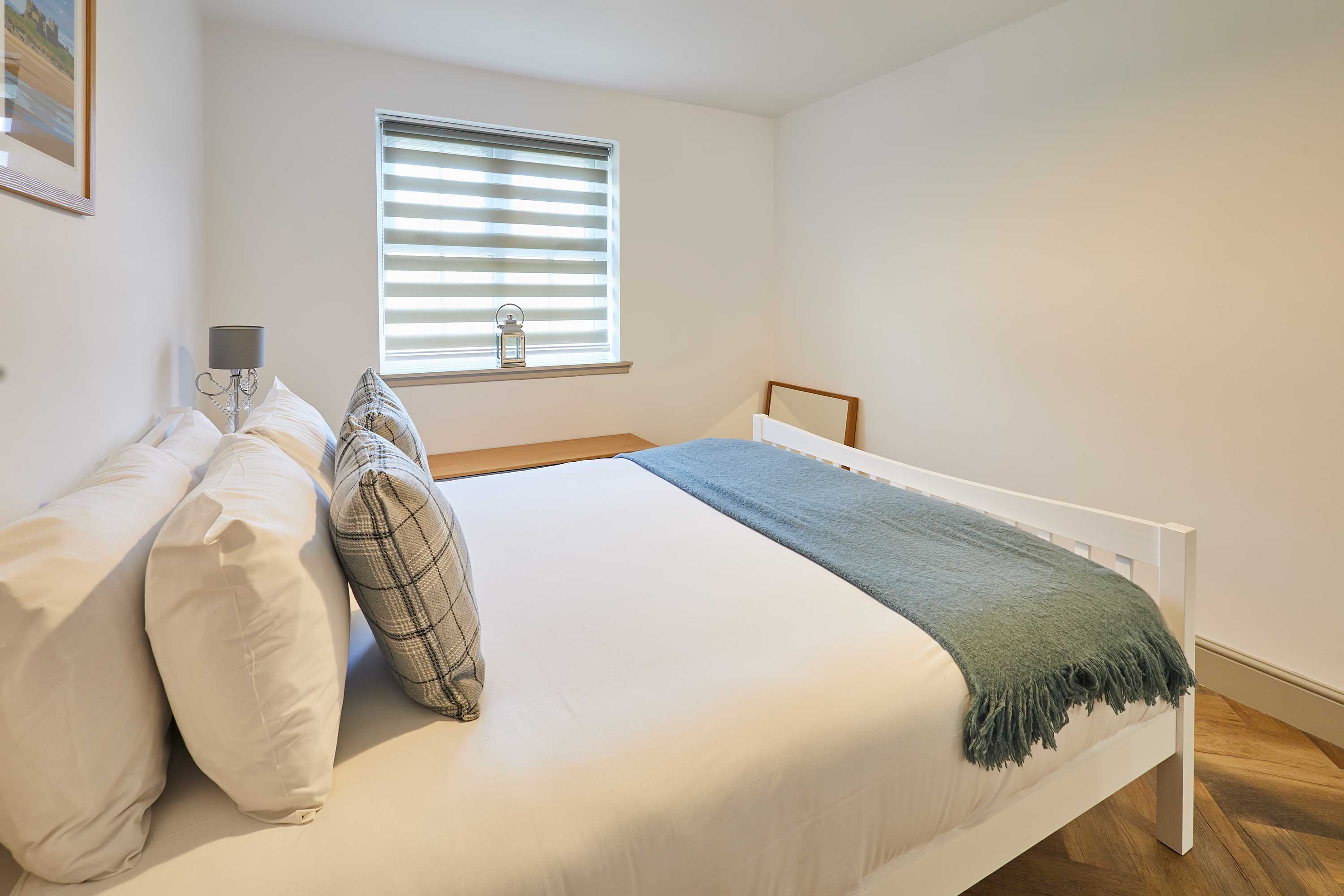Ein Schlafzimmer mit Bett, Kissen, einer Nachttischlampe, einem Fenster mit Jalousien und Holzboden.