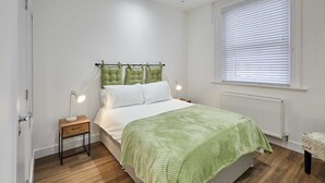 Ein ordentlich eingerichtetes Schlafzimmer mit Bett, Nachttischlampen, einem Nachttisch, einem Fenster mit Jalousien und einer Heizung.