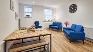 A modern living room with a wooden table, blue sofas, and a large wall clock. - Host Stay The Coach House Apartment 2 (Saltburn-by-the-Sea)