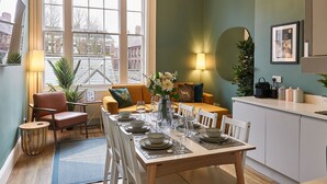 A dining area with a wooden table set for four, surrounded by white chairs. There is a window with a view of buildings outside.