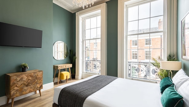 A bedroom with a large bed, a wooden dresser, a mirror, a small table, and a TV on the wall.