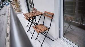 A balcony with two wooden chairs and a table, a white wall, and a view of parked cars and buildings.