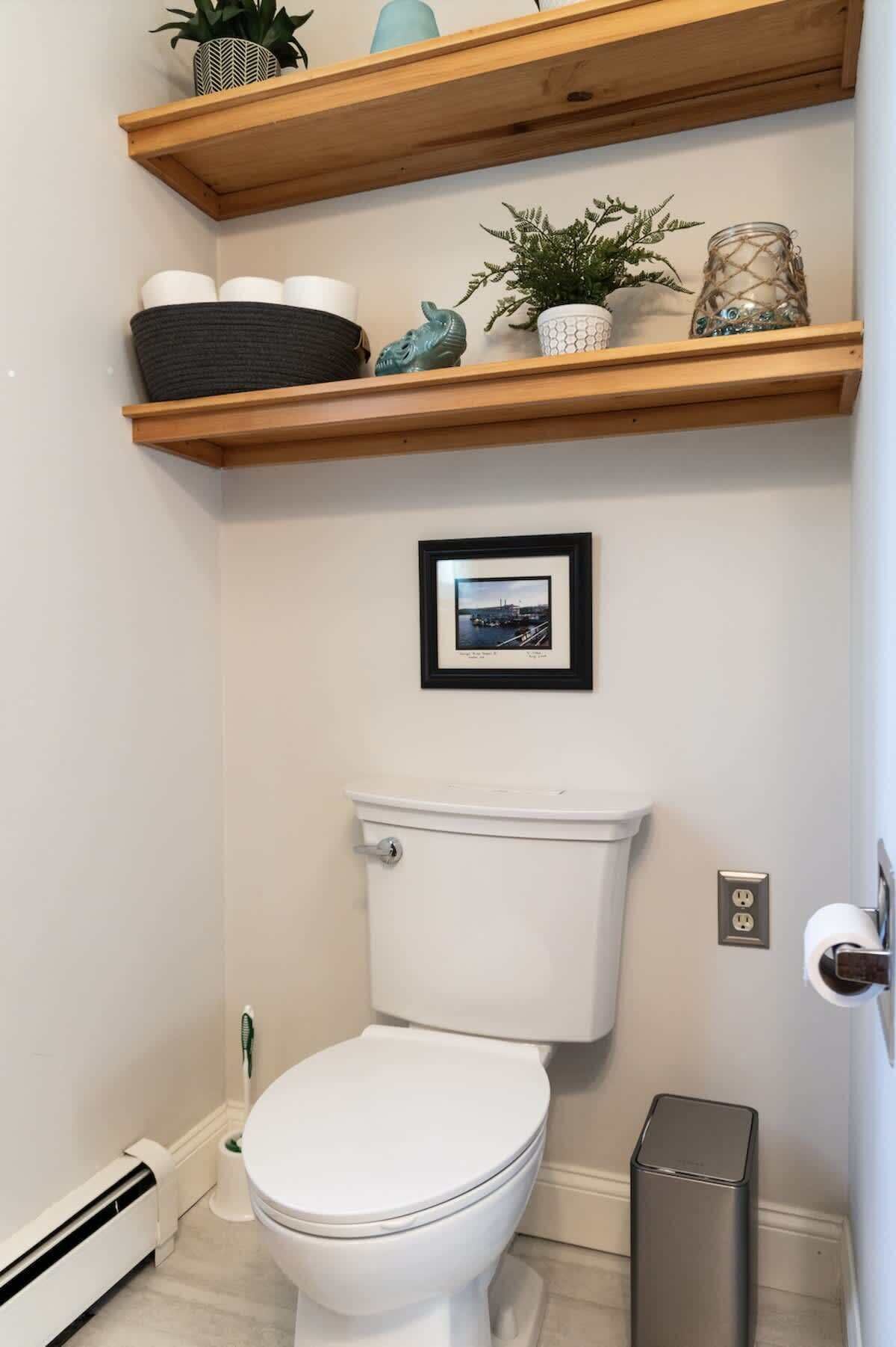 Ein Badezimmer mit einer weißen Toilette, einem gerahmten Bild an der Wand und Holzregalen mit verschiedenen Gegenständen.
