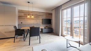 A modern living room with a dining area, a flat-screen TV mounted on a wooden wall, and large windows offering a view of the outdoors. - Amazing Apartment in Tyrol With Infrared Sauna (Westendorf)